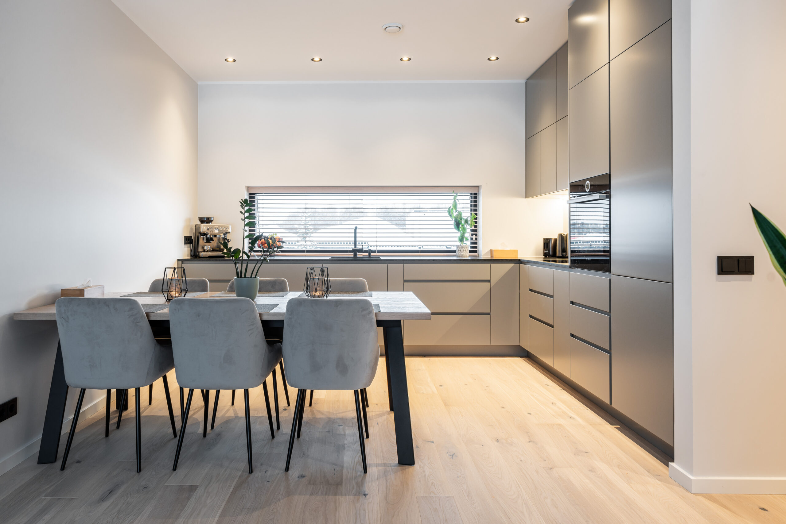 Handleless grey-beige matt fronts: a dark worktop runs beneath a ribbon window with the sink centred in it. On the right a run of tall units reaches the ceiling with a built-in oven; on the left a dining table joined to a wall-mounted shelf.